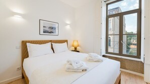 Een keurig opgemaakt bed met witte lakens, een ingelijst schilderij aan de muur, een bedlampje en een raam met uitzicht op gebouwen.