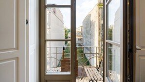 Uitzicht vanaf een balkon met een houten bank en metalen hekwerk, over een smalle straat met gebouwen en een heldere lucht.