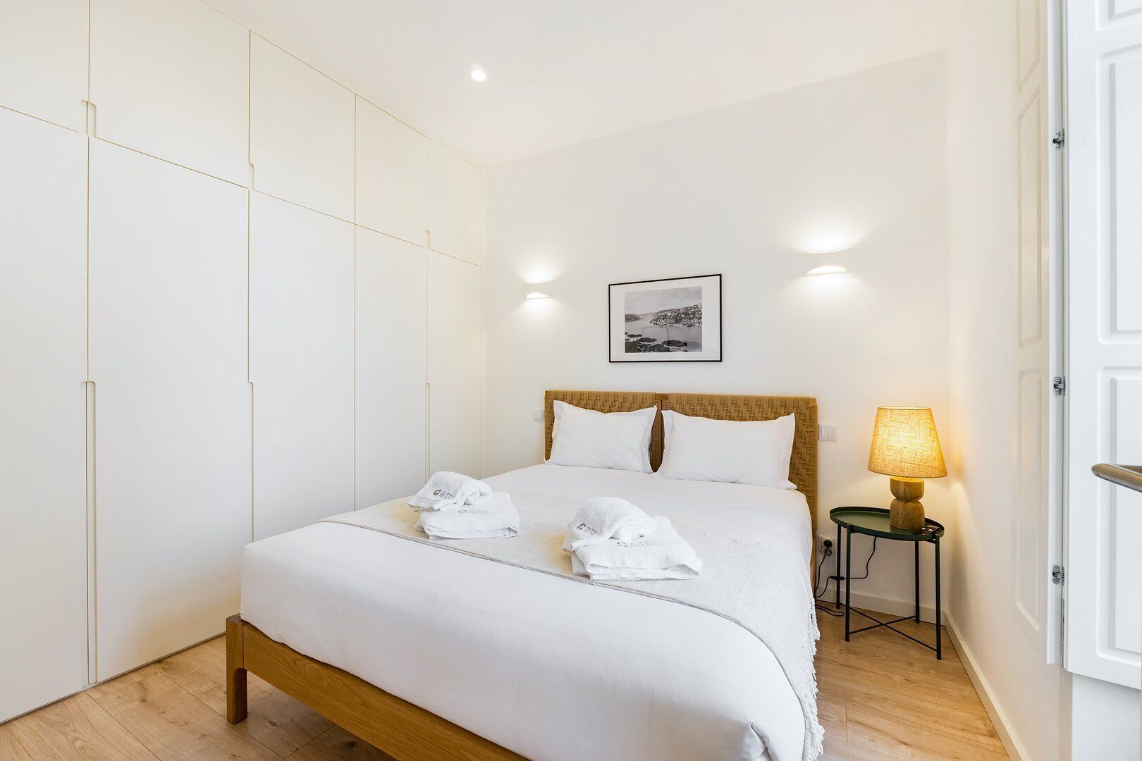 A neatly made bed with white linens and two towels, a bedside lamp, and a framed picture on the wall.