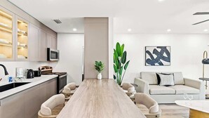 A modern kitchen with a central island, a dining area with a long table, and a living space with a sofa and a coffee table.