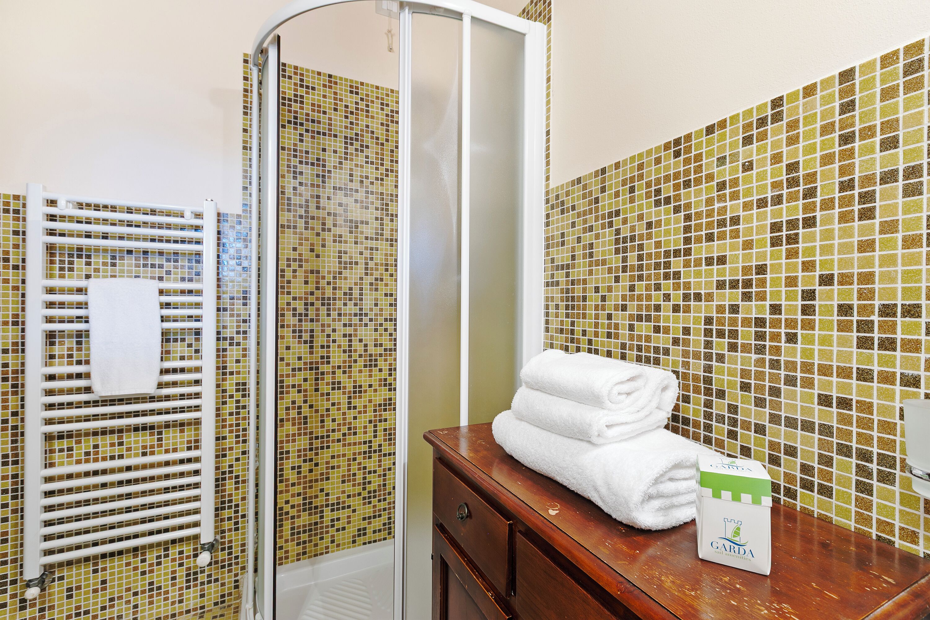 Une salle de bain avec une douche à parois de verre, un porte-serviettes blanc et un meuble-lavabo en bois sur lequel se trouve une boîte étiquetée « GARDA ».