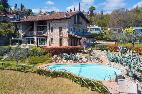 A stone villa with a swimming pool, surrounded by landscaped gardens and a view of neighboring houses.
