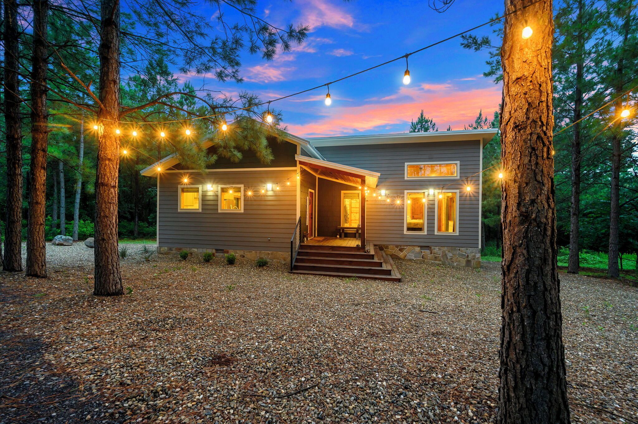 A two-story house with a porch, surrounded by trees and gravel driveway, at dusk.