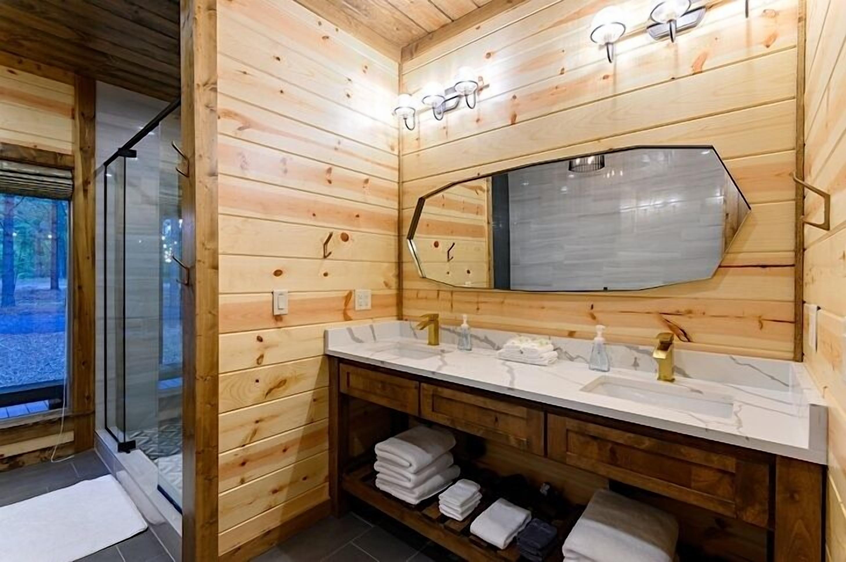 A wooden cabin bathroom with a large mirror, two sinks, and a glass shower enclosure.