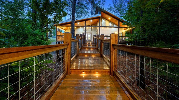 Un pont en bois mène à un chalet doté d’une porte et de fenêtres en verre, le tout étant entouré d’arbres.