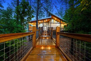 Un pont en bois mène à un chalet doté d’une porte et de fenêtres en verre, le tout étant entouré d’arbres.