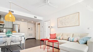 A modern living room with a sofa, two red stools, a glass table with chairs, a hanging light fixture, and a painting on the wall.