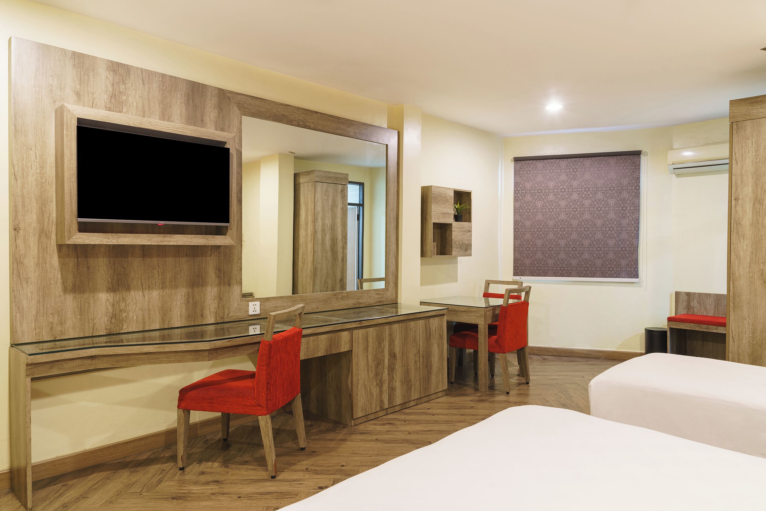 A hotel room with a wooden desk, a large mirror, a television, two red chairs, and a bed.