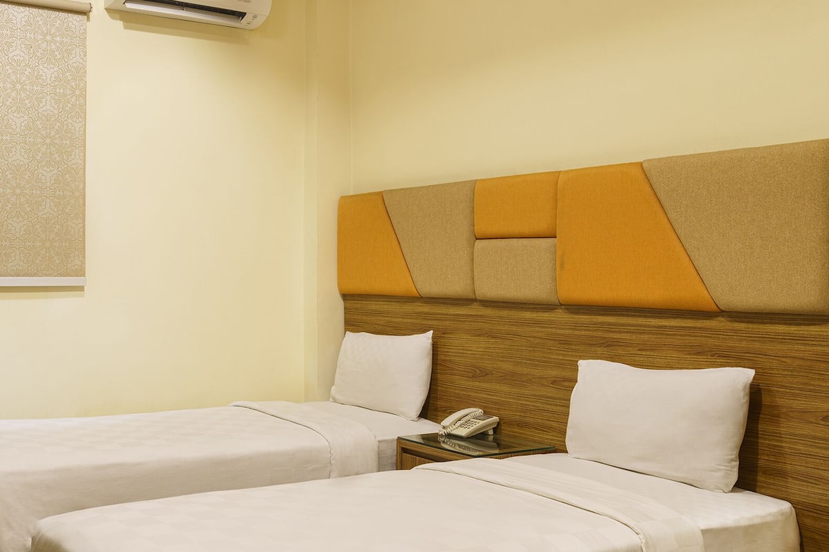 Une chambre d’hôtel avec deux lits simples, une tête de lit en bois, une table de chevet avec un téléphone et un appareil de climatisation fixé au mur.