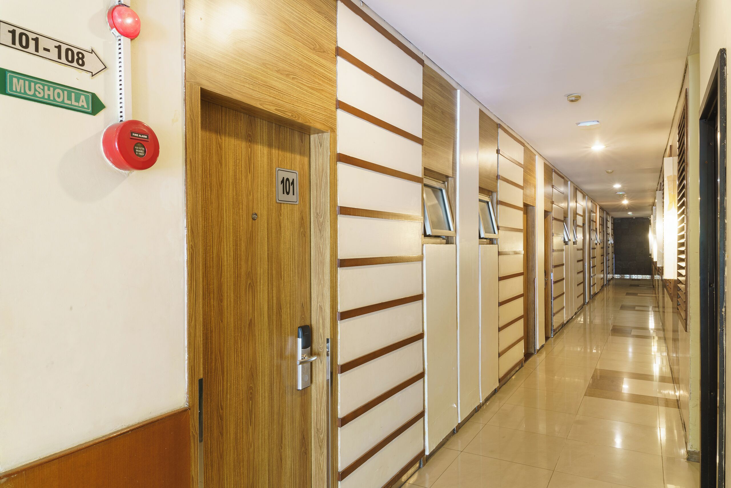 A hotel corridor with a wooden door labeled "101", a fire alarm, and a directional sign for "101-108 Musholla".