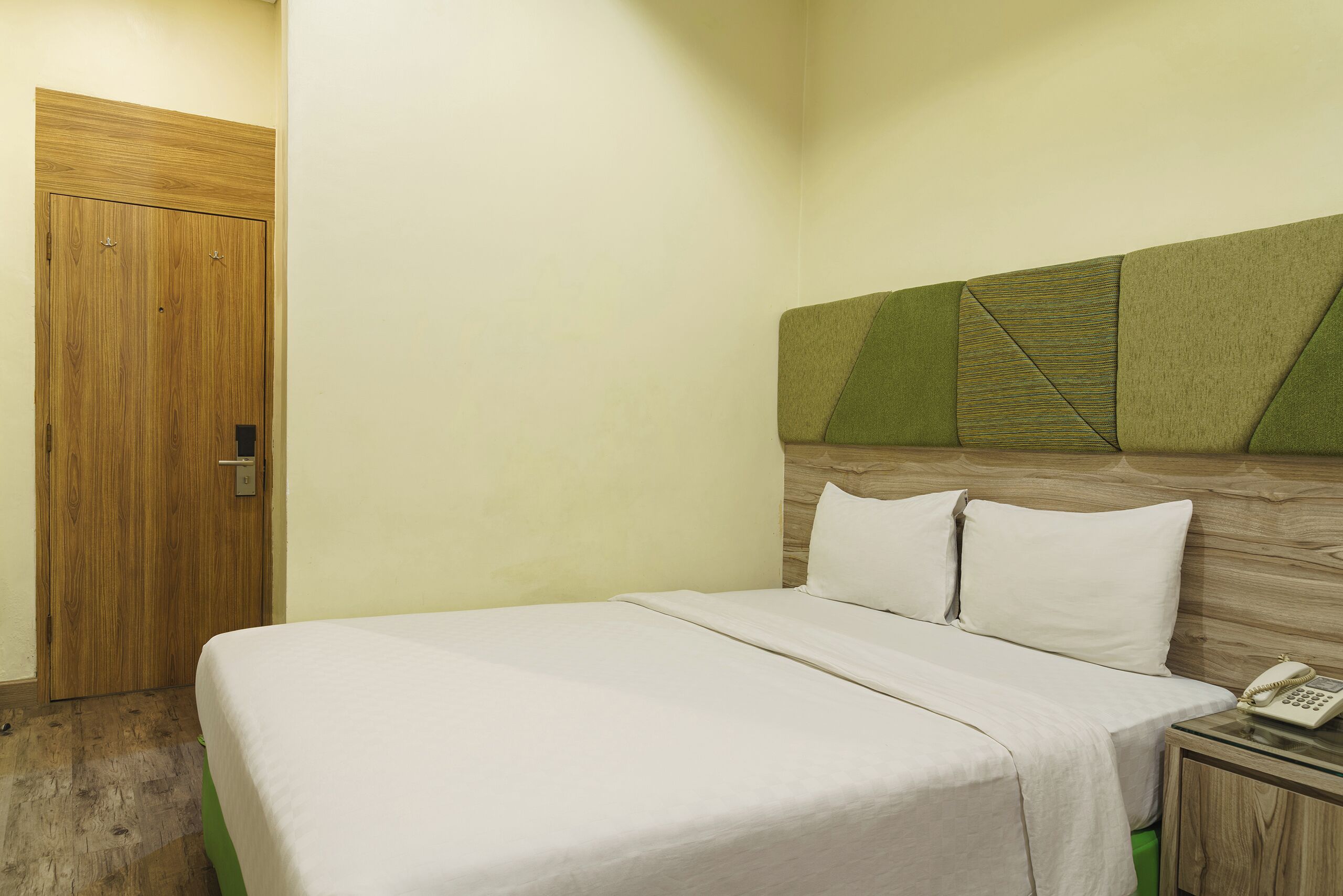 A hotel room with a wooden door, a bed with a green headboard, white bedding, and a bedside table with a telephone.
