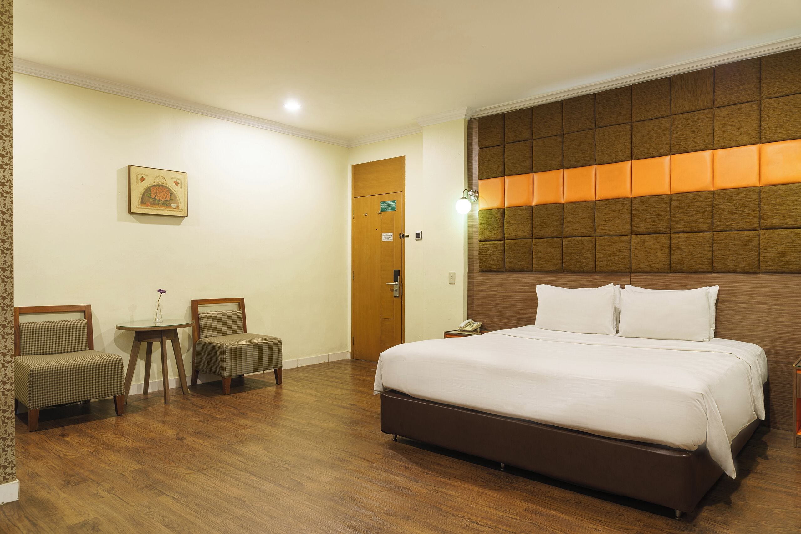 A hotel room with a large bed, two chairs, a small table, and a wooden headboard with illuminated panels.