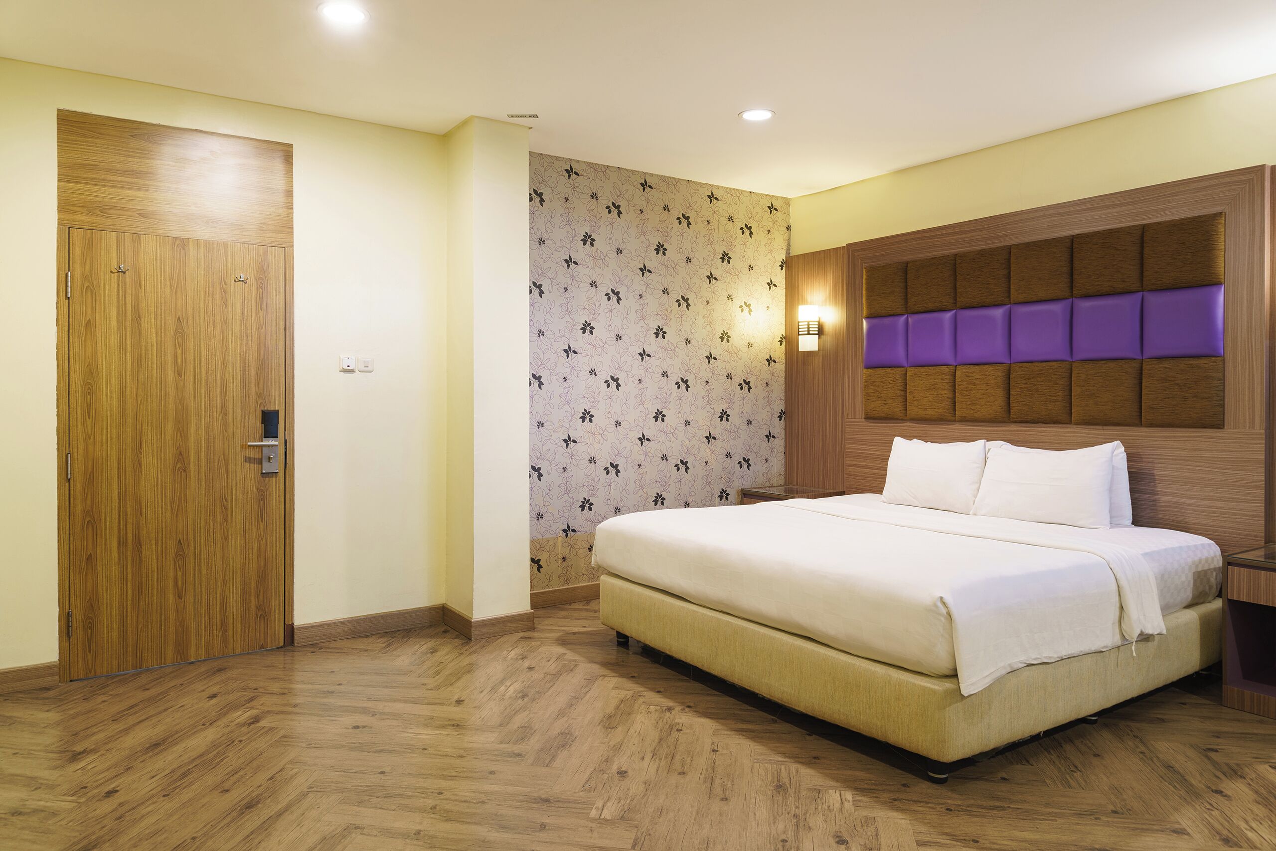 A hotel room with a wooden wardrobe, a bed with a headboard, and a patterned wallpaper.