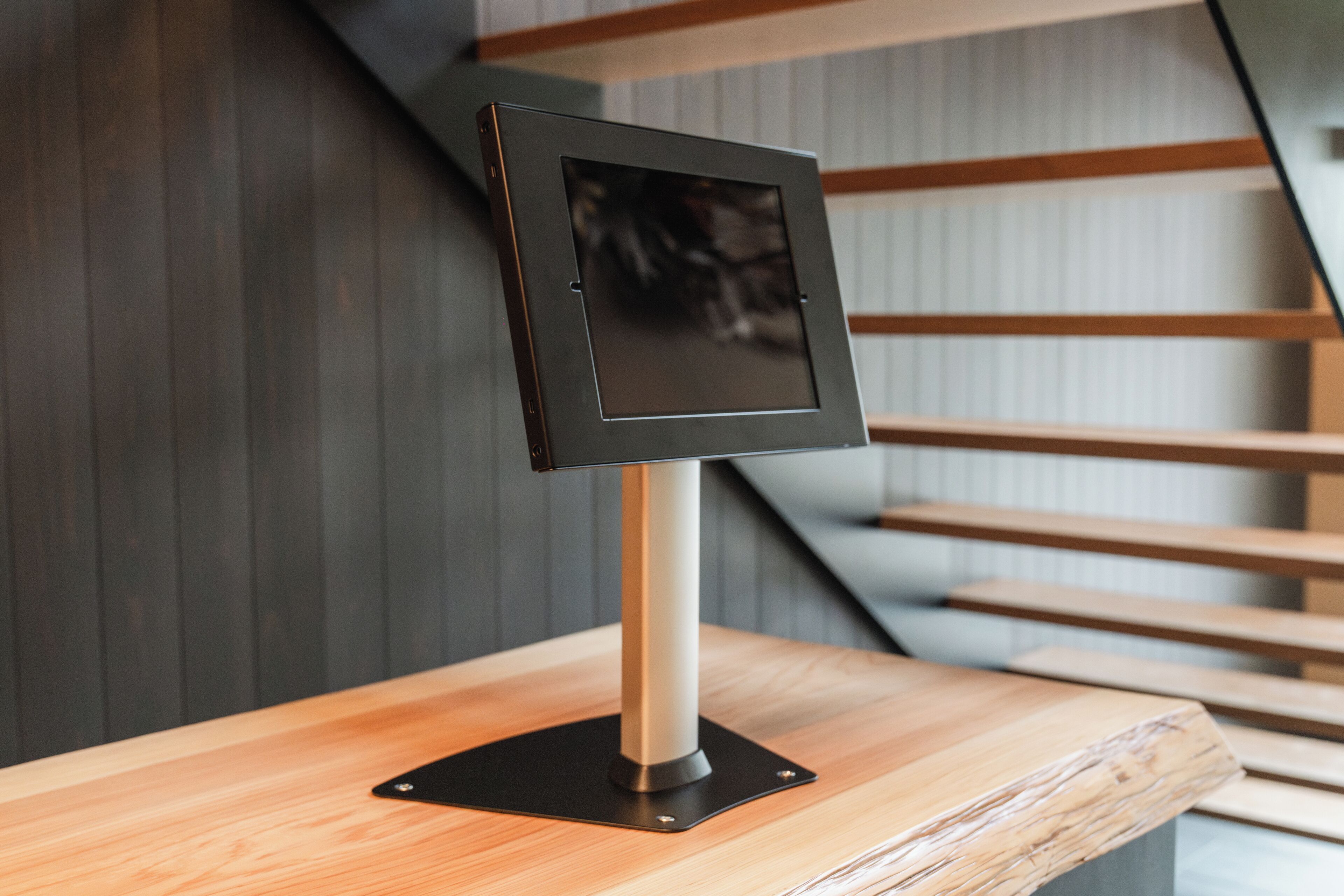 A modern, minimalist reception desk with a sleek, metallic frame and a black screen, positioned on a wooden counter with a staircase in the background.