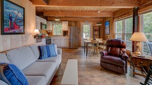 L’intérieur chaleureux d’un chalet avec un plafond en bois, un canapé agrémenté de coussins bleus, un fauteuil en cuir, une table à manger avec des chaises et un coin cuisine équipé d’un réfrigérateur et d’un four à micro-ondes.