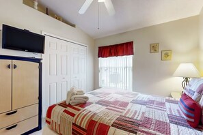 Un dormitorio con cama, televisor sobre un armario, ventana con cortinas y lámpara de noche.