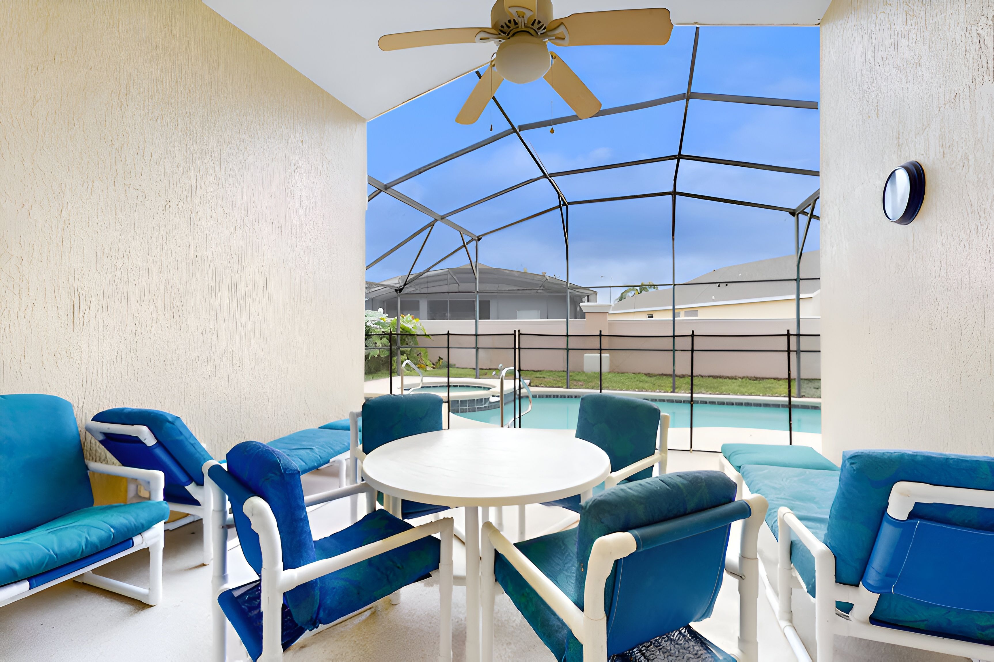 Un espace extérieur abrité, aménagé avec des meubles bleus et blancs, doté d’un ventilateur de plafond et offrant une vue sur une piscine et des bâtiments en arrière-plan.