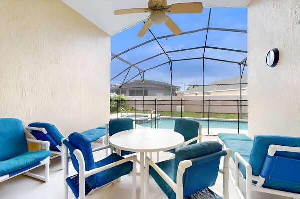 A covered outdoor seating area with blue and white furniture, a ceiling fan, and a view of a swimming pool and buildings in the background.