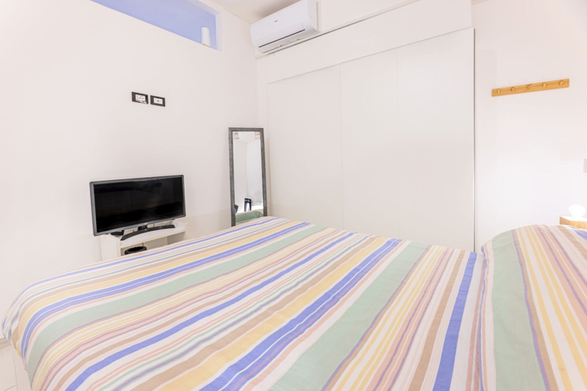 A hotel room with a bed, a television, a mirror, and a wall-mounted air conditioning unit.