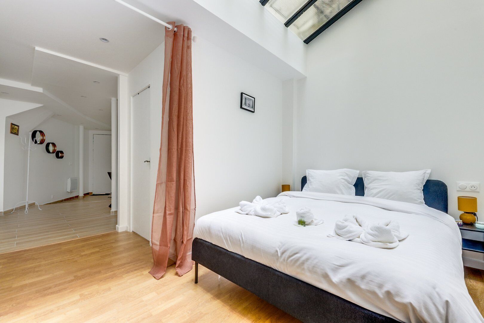 A modern hotel room with a large bed, a wooden floor, a skylight, and a view of the hallway.