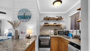 A modern kitchen with a stove, microwave, and dishwasher. There is a wooden shelf above the stove and a clock on the wall. - Stylish and modern ground-floor studio, conveniently located near 30A! (Panama City Beach)