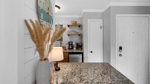 A kitchen with a granite countertop, a wall-mounted clock, a vase with pampas grass, and a coffee maker. - Stylish and modern ground-floor studio, conveniently located near 30A! (Panama City Beach)