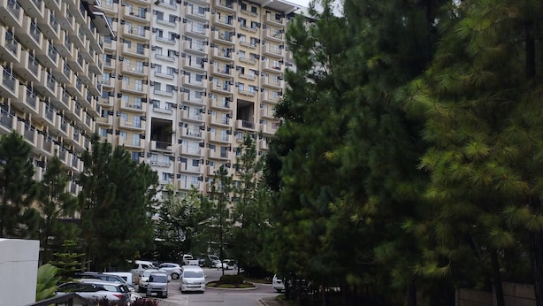 A multi-story residential building with balconies, a parking area with cars, and surrounding trees.