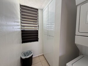 A small, compact bathroom with a toilet, a trash bin, and a window with blinds.