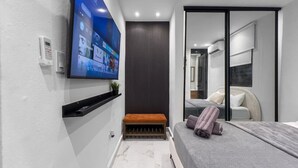Una moderna camera d'hotel con una grande TV a parete, una panca, un letto con asciugamani e una porta scorrevole in vetro che dà accesso al bagno.
