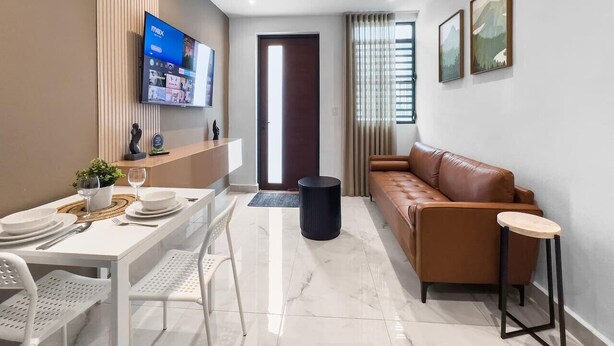 Un soggiorno moderno con un divano in pelle marrone, un tavolo da pranzo apparecchiato con piatti e bicchieri bianchi, una TV a parete e una porta che dà su un'altra stanza.
