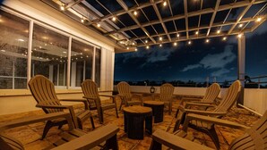 Un espace extérieur aménagé avec des chaises Adirondack et une table ronde, situé sous une terrasse couverte et illuminée de guirlandes lumineuses.