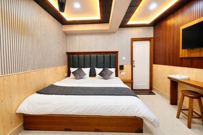 Ein Schlafzimmer mit einem Holz-Kopfteil, einem Bett mit weißer Bettwäsche, einem Schreibtisch mit Stuhl und einem an der Wand befestigten Fernseher.