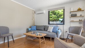 Ein modernes Wohnzimmer mit grauem Sofa, zwei Sesseln, einem Holztisch und einem großen Fenster mit Blick auf Grünflächen.