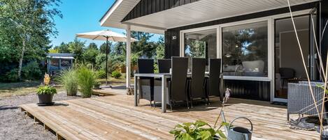 Una terraza de madera con mobiliario exterior, una pérgola y vistas al interior.