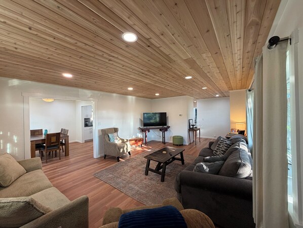 A living room with a wooden ceiling, a TV, a sofa, a coffee table, and a dining area with a table and chairs.