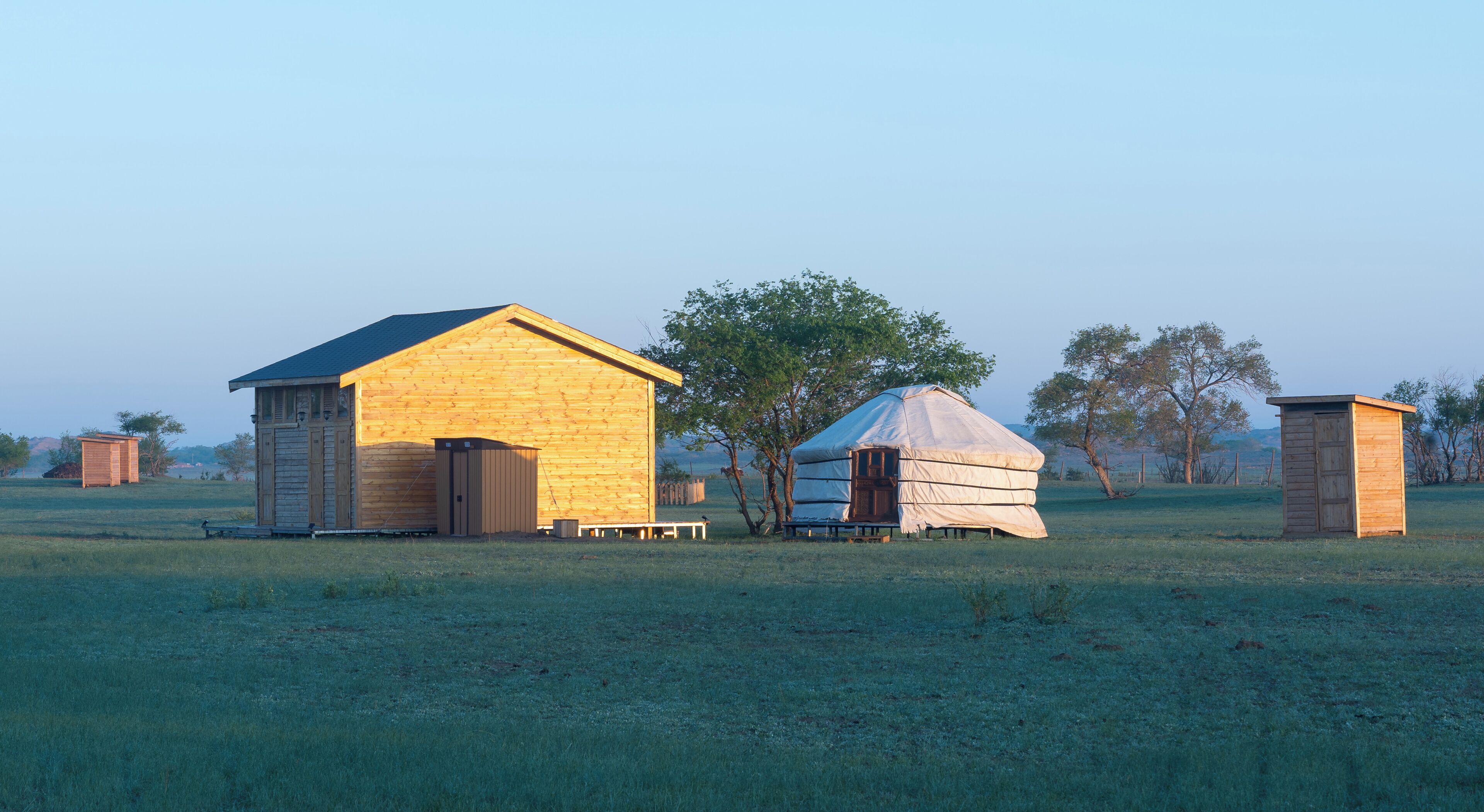 A rustic wooden building with a sloped roof, a small wooden structure with a flat roof, and a large tent-like structure in a grassy field.