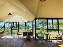 Una amplia veranda con piso de madera, ventanas grandes que ofrecen vistas de la vegetación circundante, y varias sillas y mesas para comer o descansar.