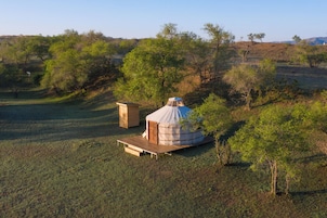 Una yurta con terraza de madera y una pequeña construcción de madera al lado, ubicada en un campo cubierto de hierba y árboles.