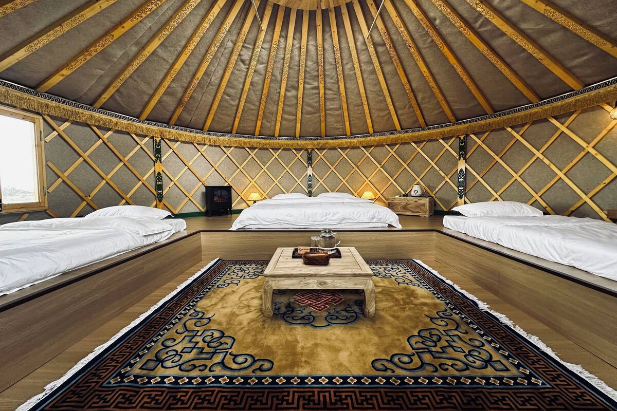 A traditional yurt interior with three beds, a central wooden table, and a patterned rug.