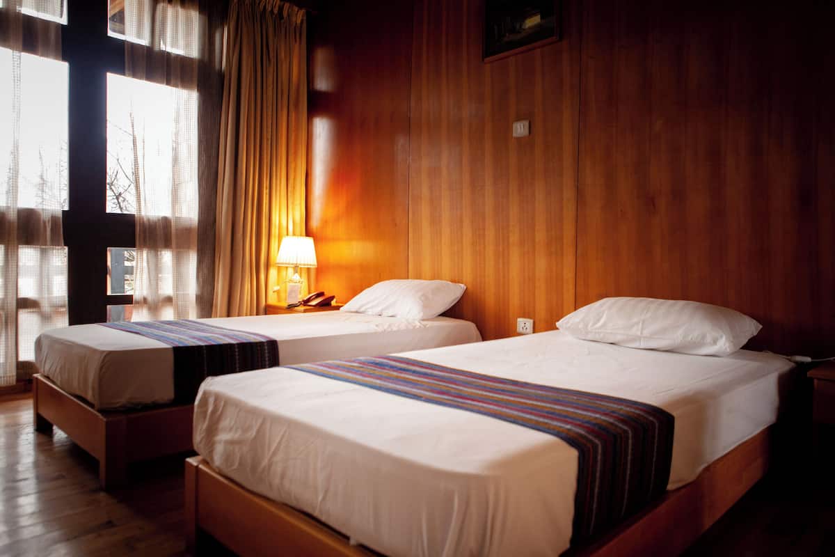 A hotel room with two single beds, wooden walls, a window with curtains, and a bedside lamp.