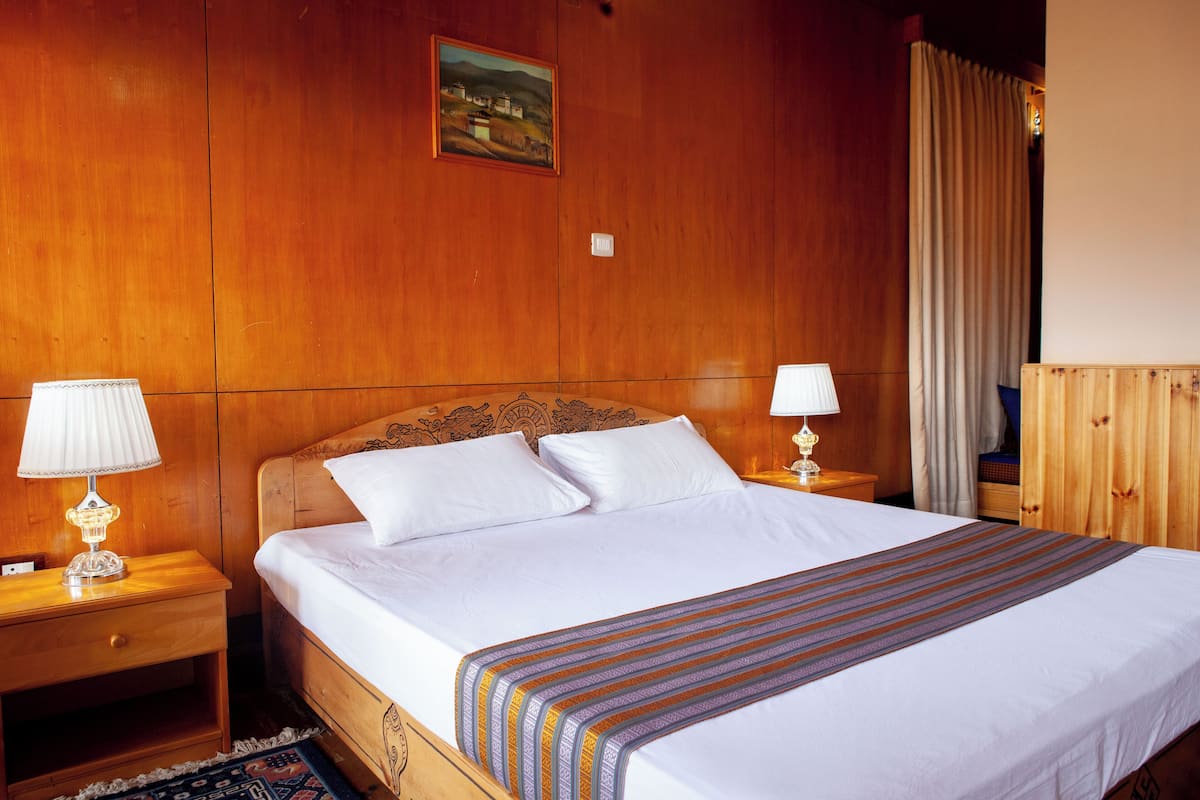 A hotel room with a wooden headboard, a large bed with white linens, two bedside tables with lamps, and a painting on the wall.