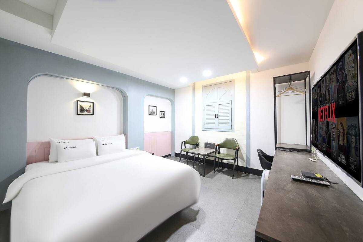 A modern hotel room with a large bed, two chairs, a small table, and a TV displaying movie posters.