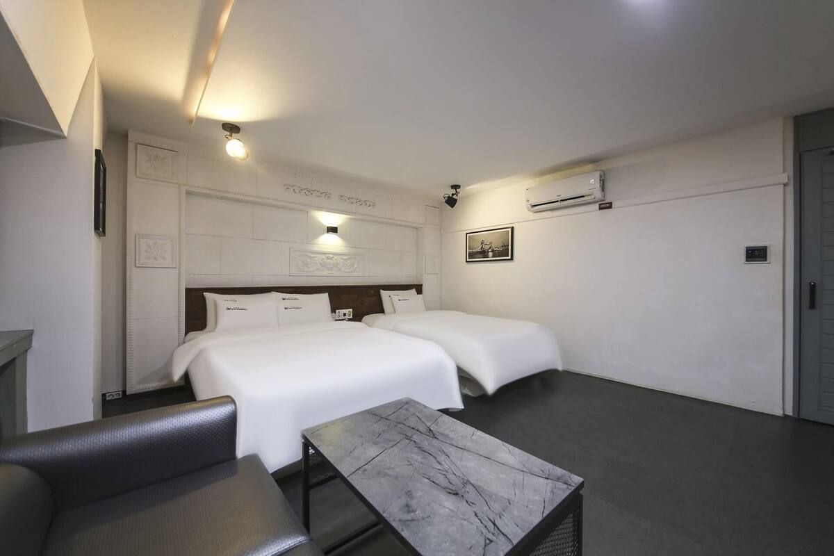 A hotel room with two beds, a small table, a chair, and a wall-mounted air conditioner.