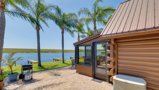 A patio with a view of a lake, palm trees, and a building with a sliding glass door. - Boat, Fish & Swim: Lakefront Cabin in Okeechobee! (Okeechobee)