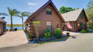 A log cabin with a view of a lake, a paved driveway, and a small garden with potted plants and flowers. - Boat, Fish & Swim: Lakefront Cabin in Okeechobee! (Okeechobee)