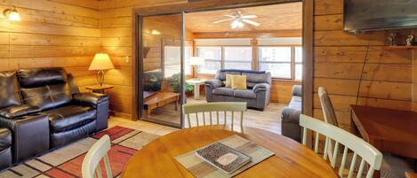 A cozy cabin interior with a wooden ceiling, a leather sofa, a dining table with chairs, and a television mounted on the wall.