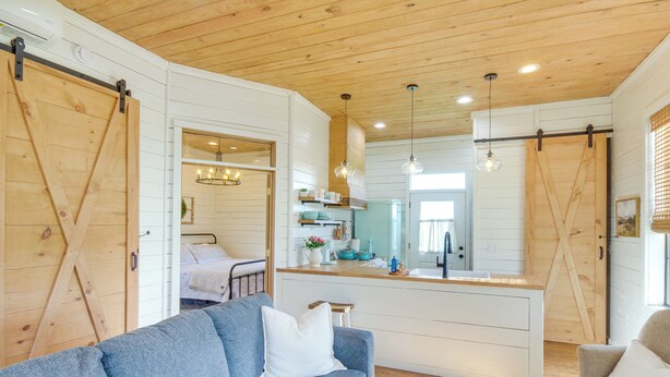 A cozy living room with a blue sofa, a wooden ceiling, and a kitchen area visible through an open door. - Contrell Cottage On Farm w/ Peaceful Views (Forest)