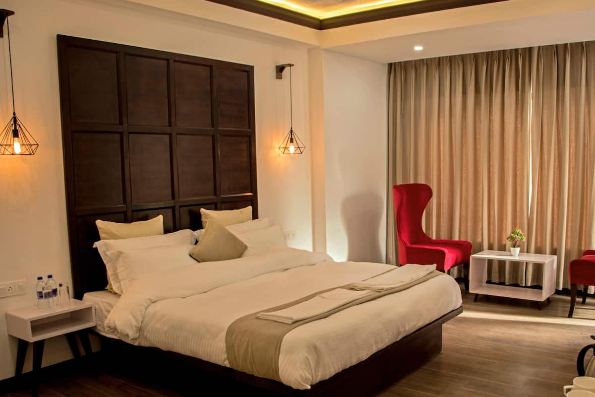 Una habitación de hotel con una cama grande, dos mesitas de noche con lámparas, un sillón rojo, una pequeña mesa con una planta y un amplio ventanal con cortinas.