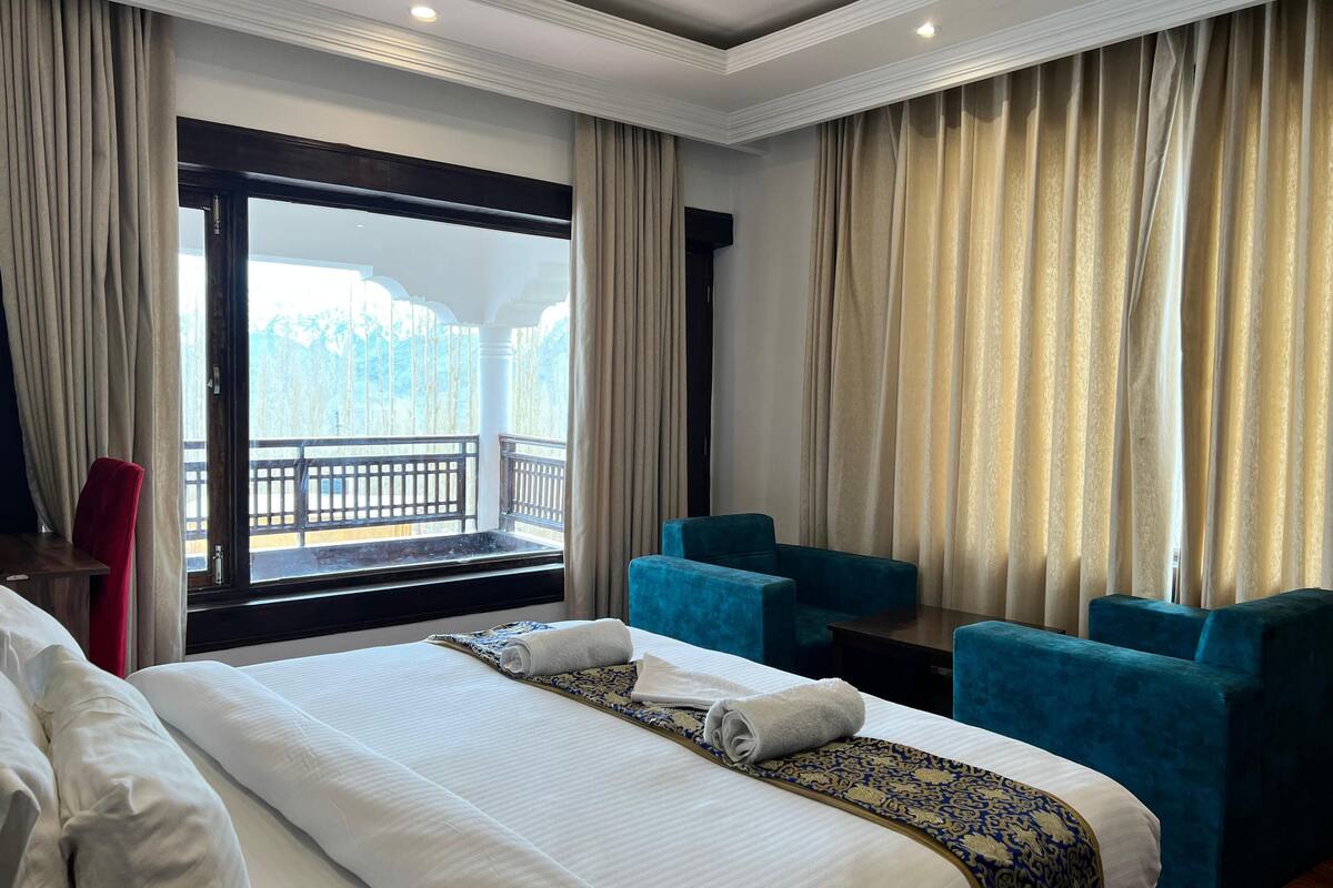 Una habitación de hotel con una cama grande, dos sillones azules, una mesita y un balcón con barandilla.