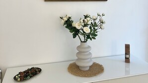 Un cuadro enmarco con un atardecer sobre el agua, un bote, un jarrón blanco con flores y un adorno pequeño sobre un tocador blanco.